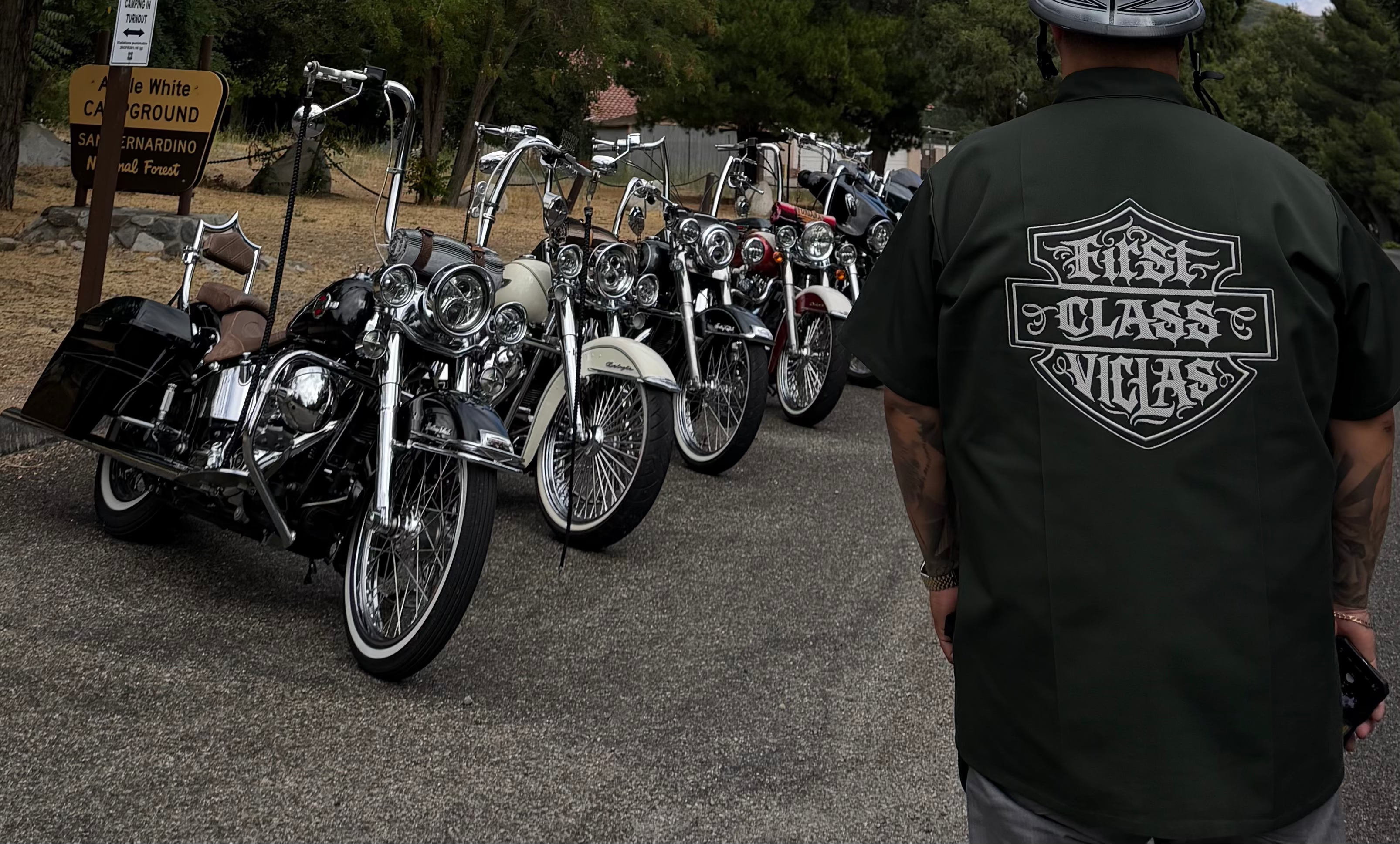 A man wearing a black graphic t-shirt with a 'Rits Class Vitas' logo on the back stands next to a lineup of classic motorcycles outdoors under a cloudy blue sky.